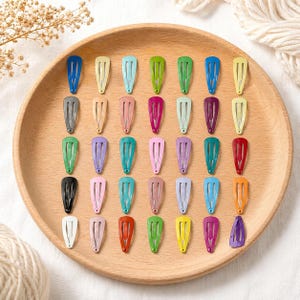 Può includere: Una collezione di fermagli per capelli colorati disposti su un vassoio di legno. I fermagli sono in varie tonalità, tra cui blu, verde, rosa, viola e giallo. I fermagli sono in metallo e hanno un design semplice ed elegante. Il vassoio è rotondo e in legno chiaro.