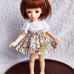 May include: A small doll with brown hair wearing a white dress with a floral print and a small embroidered bear on the bodice. The doll is standing on a brown surface.