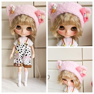 May include: A pink fuzzy doll hat with bear ears and a small ice cream cone applique. The hat is worn by a doll with blonde hair and large blue eyes. The doll is wearing a white shirt and black and white overalls.