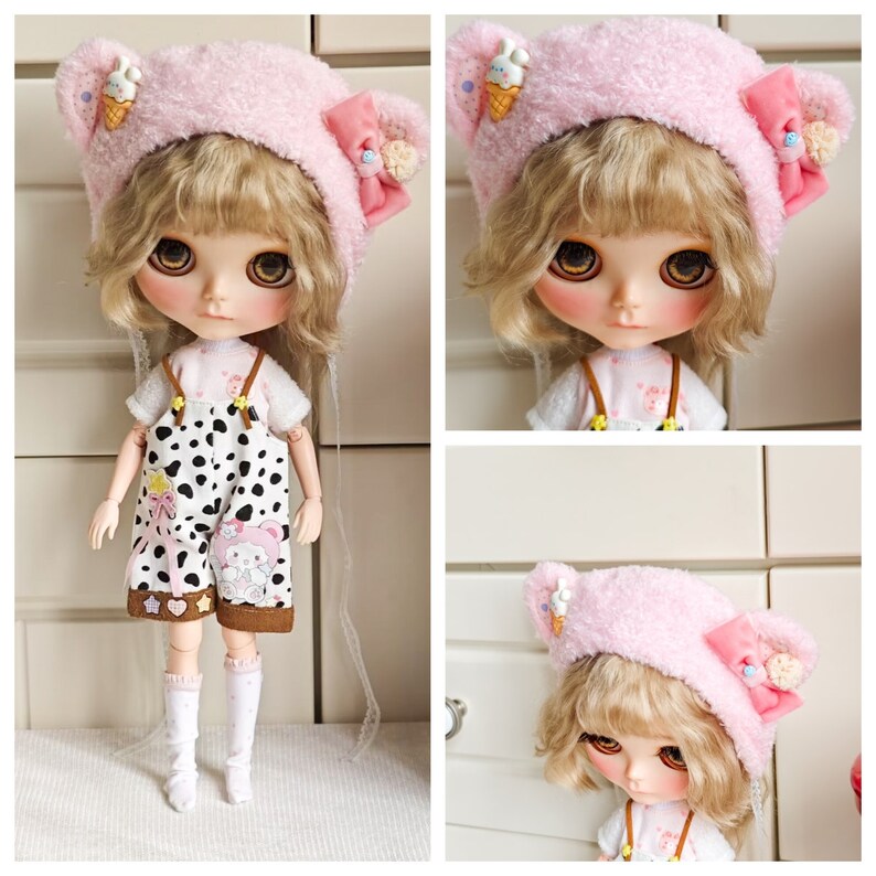 May include: A pink fuzzy doll hat with bear ears and a small ice cream cone applique. The hat is worn by a doll with blonde hair and large blue eyes. The doll is wearing a white shirt and black and white overalls.