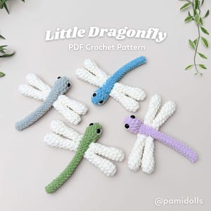 May include: Four crocheted dragonflies in blue, green, grey, and purple with white wings. Each dragonfly has black eyes. The text "Little Dragonfly PDF Crochet Pattern" is at the top of the image.