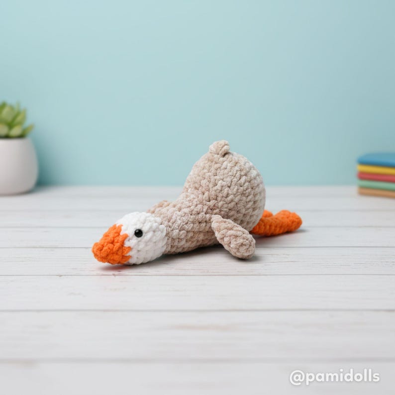 May include: A handmade crochet goose doll in a lying down position. The goose has a beige body, white head, and an orange beak and feet. The doll has a black eye and is lying on a white wooden surface.
