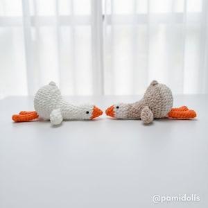 May include: Two handmade crochet geese in white and tan, with orange beaks and feet, are lying on a white surface. The geese are facing each other, with a soft, textured appearance. The background is a blurred white curtain.