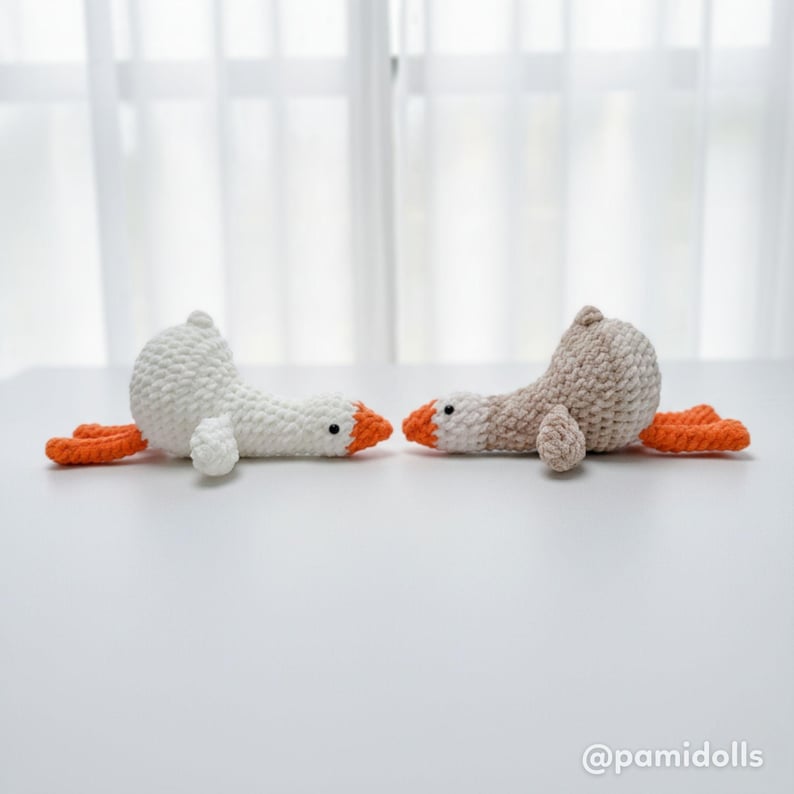 May include: Two handmade crochet geese in white and tan, with orange beaks and feet, are lying on a white surface. The geese are facing each other, with a soft, textured appearance. The background is a blurred white curtain.