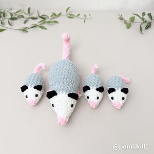 K&ouml;nnte beinhalten: Geh&auml;kelte Opossum-Familie in Grau, Wei&szlig;, Schwarz und Rosa. Die Opossums haben schwarze Ohren, rosa Nasen und rosa Schw&auml;nze. Das gr&ouml;&szlig;te Opossum liegt, w&auml;hrend die kleineren aufrecht stehen. Die Familie ist auf einer hellgrauen Oberfl&auml;che angeordnet.