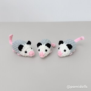 K&ouml;nnte beinhalten: Drei geh&auml;kelte Opossums. Jedes hat einen grauen K&ouml;rper, ein wei&szlig;es Gesicht, schwarze Ohren und eine rosa Nase und einen rosa Schwanz. Die Opossums sind handgefertigt und wirken weich und kuschelig. Der Hintergrund ist einfarbig hellgrau.
