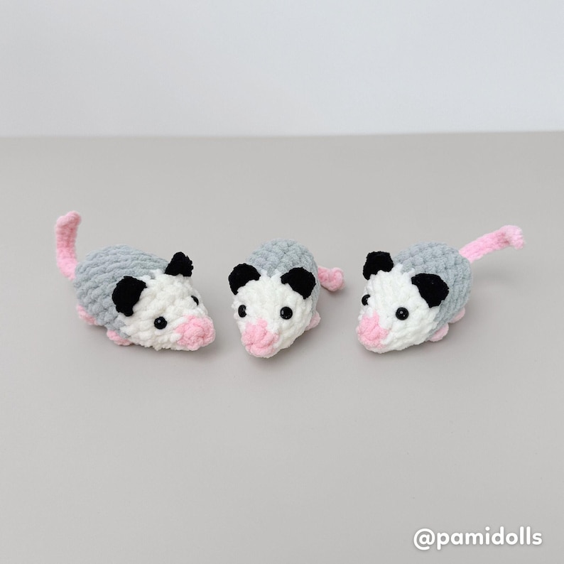 K&ouml;nnte beinhalten: Drei geh&auml;kelte Opossums. Jedes hat einen grauen K&ouml;rper, ein wei&szlig;es Gesicht, schwarze Ohren und eine rosa Nase und einen rosa Schwanz. Die Opossums sind handgefertigt und wirken weich und kuschelig. Der Hintergrund ist einfarbig hellgrau.