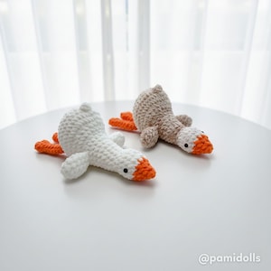 May include: Two handmade crochet geese toys. One is white with an orange beak and feet, the other is tan with an orange beak and feet. The geese are lying down on a white surface.