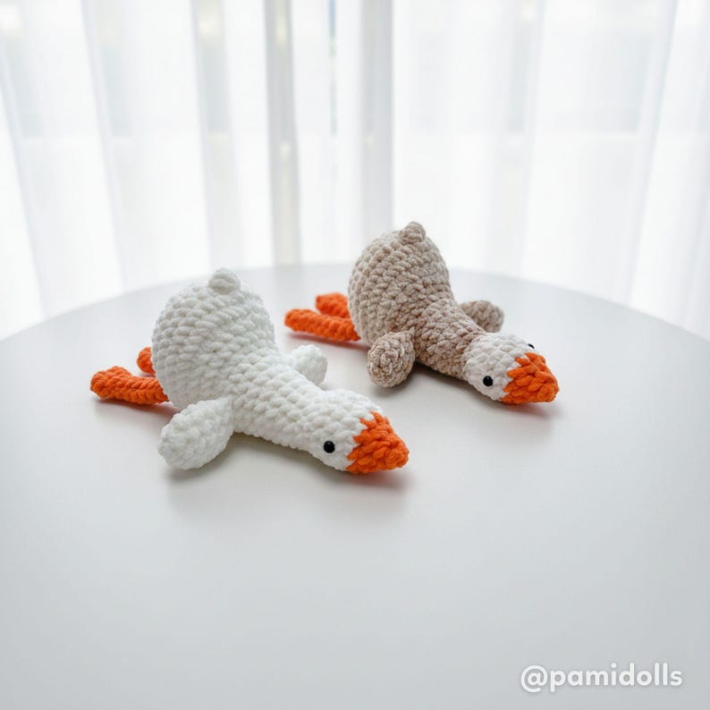 May include: Two handmade crochet geese toys. One is white with an orange beak and feet, the other is tan with an orange beak and feet. The geese are lying down on a white surface.