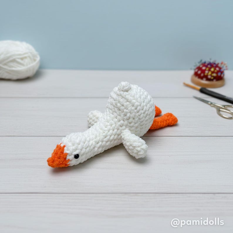 May include: A handmade white crochet goose with an orange beak and feet. The goose is lying on its back on a white wooden surface. The background is a light blue wall. A ball of white yarn, a pincushion, and scissors are also visible.