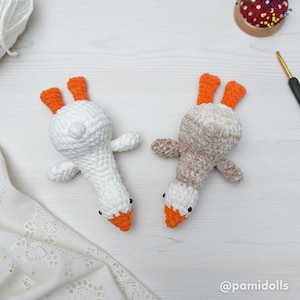 May include: Two handmade crochet goose toys. One is white with orange feet and beak, the other is beige with white and orange accents. Both have black eyes. The toys are lying on a white surface.