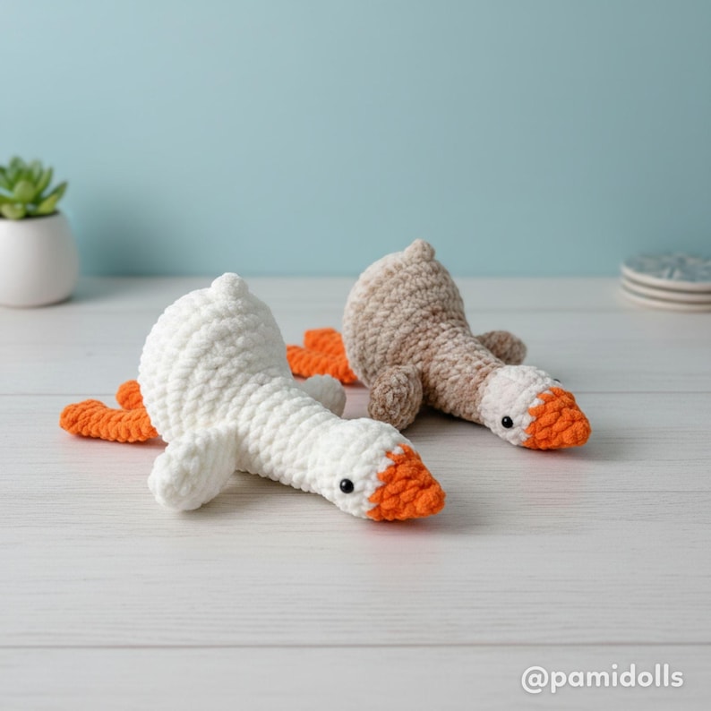 May include: Two handmade crochet geese, one white and one tan, with orange beaks and feet, lie on a light-colored surface. The geese have black eyes and are positioned as if resting. The background is a light blue wall.