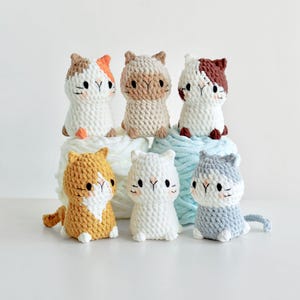 May include: Six crocheted cat toys in various colours, including white, brown, orange, and grey. The cats are sitting on a pile of yarn.