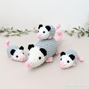 K&ouml;nnte beinhalten: Eine Sammlung von vier geh&auml;kelten Opossum-Pl&uuml;schtieren. Die Opossums haben graue K&ouml;rper, wei&szlig;e Gesichter, schwarze Ohren und rosa Nasen, F&uuml;&szlig;e und Schw&auml;nze. Das gr&ouml;&szlig;te Opossum hat ein kleineres oben drauf. Die Pl&uuml;schtiere sind handgefertigt.