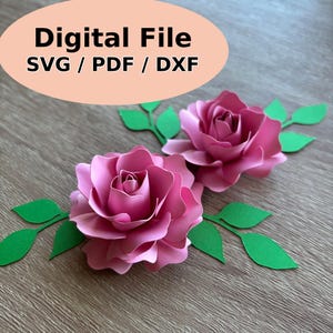 May include: Two pink paper roses with green paper leaves are arranged on a wooden surface. The roses are in full bloom, with detailed petal layers. The text "Digital File SVG / PDF / DXF" is in a peach-colored oval in the upper left.