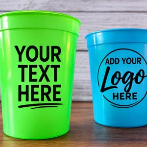 May include: Two plastic stadium cups, one green and one blue, are displayed on a wooden surface. The green cup has the text "YOUR TEXT HERE" in black. The blue cup has the text "ADD YOUR Logo HERE" in black.