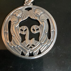 May include: A silver pendant with a circular design. The pendant features two angels with outstretched wings, framing a central design. The angels are detailed with facial features and flowing hair. The pendant has a loop at the top for hanging.