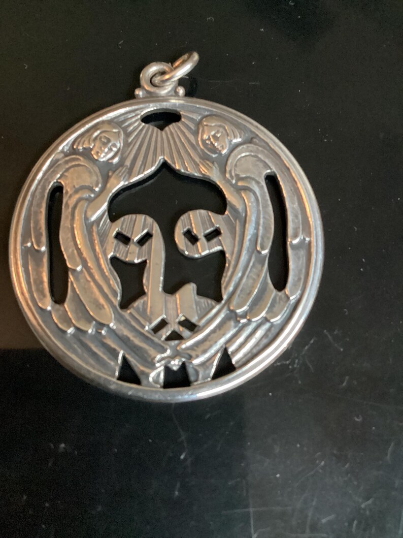 May include: A silver pendant with a circular design. The pendant features two angels with outstretched wings, framing a central design. The angels are detailed with facial features and flowing hair. The pendant has a loop at the top for hanging.