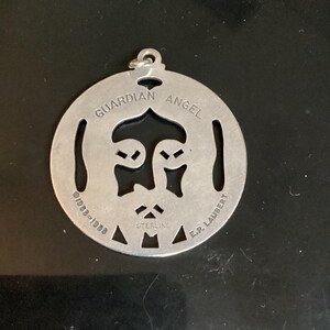 May include: A circular, silver-toned pendant featuring the words "GUARDIAN ANGEL" and a face design. The pendant is marked "STERLING" and "E.P. LAUBERT" with the date "1939-1988".