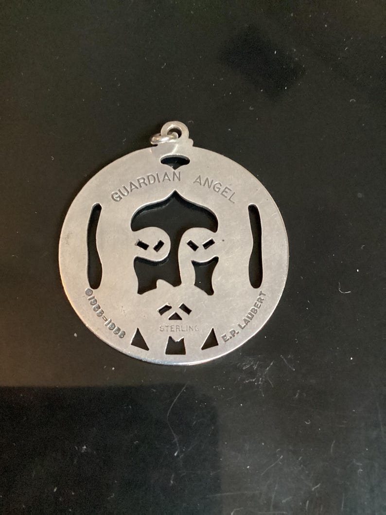 May include: A circular, silver-toned pendant featuring the words "GUARDIAN ANGEL" and a face design. The pendant is marked "STERLING" and "E.P. LAUBERT" with the date "1939-1988".