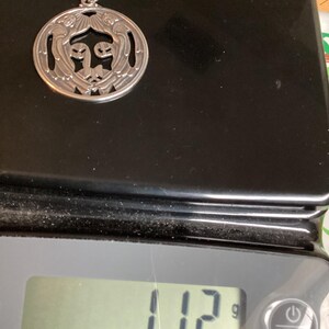 May include: A silver pendant with a circular design featuring stylized figures and sunburst details. The pendant weighs 11.2 grams. The pendant is on a black surface.