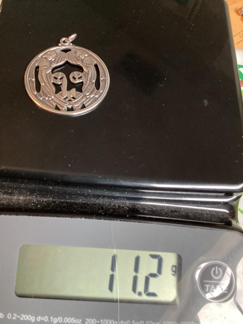 May include: A silver pendant with a circular design featuring stylized figures and sunburst details. The pendant weighs 11.2 grams. The pendant is on a black surface.