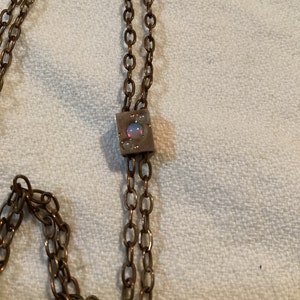 May include: A bronze-toned necklace with a square pendant featuring an iridescent stone. The necklace has a double-strand chain design, with a simple, elegant aesthetic. The pendant is the focal point, adding a touch of color and interest to the piece.