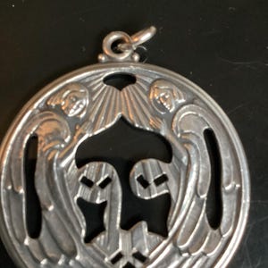 May include: A silver pendant with an intricate design featuring two figures with wings and a central stylized shape. The pendant has a loop at the top for a chain. The design is detailed and the metal has a polished finish.