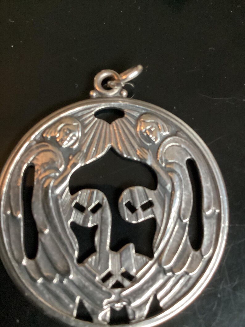May include: A silver pendant with an intricate design featuring two figures with wings and a central stylized shape. The pendant has a loop at the top for a chain. The design is detailed and the metal has a polished finish.