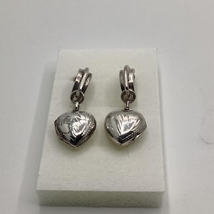 May include: A pair of silver-toned heart-shaped earrings. Each earring features a detailed design on the heart pendant and a decorative top. The earrings are displayed on a white surface against a white background.