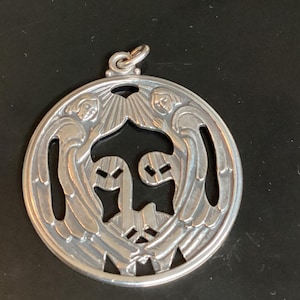 May include: A silver pendant with a circular design. The pendant features two angels with outstretched wings and a central design. The pendant has a loop at the top for a chain.