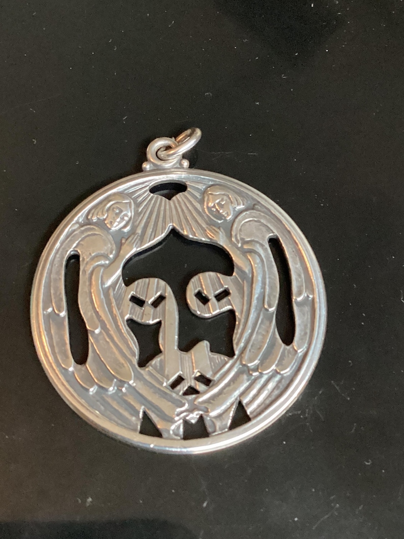 May include: A silver pendant with a circular design. The pendant features two angels with outstretched wings and a central design. The pendant has a loop at the top for a chain.