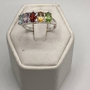 May include: A silver ring featuring five oval-cut gemstones in a row. The gemstones are light blue, purple, red, yellow, and green. The ring is displayed on a white, hexagonal stand.