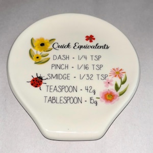 May include: White ceramic spoon rest with floral and ladybug illustrations. The spoon rest has the text "Quick Equivalents" and measurements for dash, pinch, smidge, teaspoon (42g), and tablespoon (15g).