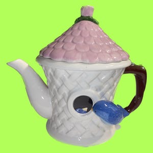 May include: A white ceramic teapot in the shape of a birdhouse, with a pink roof and a blue bird on the side. The teapot has a brown handle and a white spout. The birdhouse design includes a round opening.