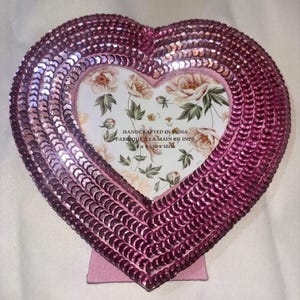 Könnte beinhalten: Ein herzförmiger Fotorahmen mit rosa Pailletten. Der Rahmen hat einen floralen Hintergrund und einen Ständer. Der Text "HANDCRAFTED IN INDIA FABRIQUE A LA MAIN EN INDE x 10 x 10cm" ist auf dem Rahmen aufgedruckt.