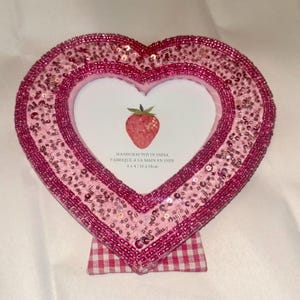 Könnte beinhalten: Rosa herzförmiger Bilderrahmen, verziert mit Pailletten und Perlen. Der Rahmen hat einen weißen Innenraum mit einer Erdbeer-Illustration. Der Rahmen wird von einem Vichy-Karo-Sockel getragen. Rahmenabmessungen: 10 x 10 cm.