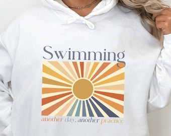 Op de afbeelding: Witte hoodie met het woord "Swimming" boven een retro graphic van een zonnestraalontwerp in tinten geel, oranje en blauw. De tekst "another day, another practice" staat eronder. De hoodie heeft een capuchon met trekkoord.