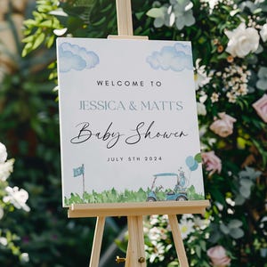 May include: A white sign with blue clouds and a golf cart illustration. The sign reads "Welcome to Jessica & Matt's Baby Shower July 5th 2024".