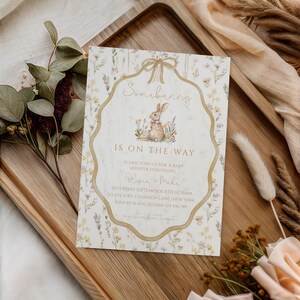 May include: A baby shower invitation with a watercolor illustration of a bunny and floral accents. The text "Somebunny is on the way" is displayed in a decorative frame. The invitation is on a wooden tray with floral and fabric accents.
