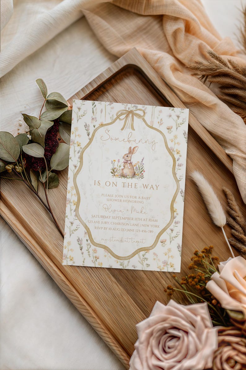 May include: A baby shower invitation with a watercolor illustration of a bunny and floral accents. The text "Somebunny is on the way" is displayed in a decorative frame. The invitation is on a wooden tray with floral and fabric accents.