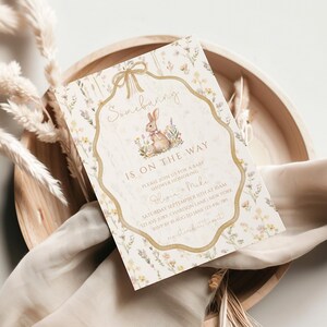 May include: A baby shower invitation with a floral design and a bunny illustration. The text "Somebunny is on the way" is displayed in elegant script. The invitation is set on a wooden tray with dried plants and a soft cloth.