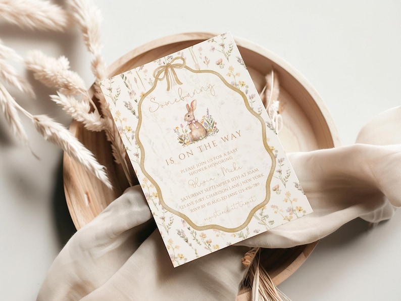 May include: A baby shower invitation with a floral design and a bunny illustration. The text "Somebunny is on the way" is displayed in elegant script. The invitation is set on a wooden tray with dried plants and a soft cloth.