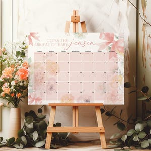 Puede incluir: Un calendario floral rosa y blanco con el texto "Guess the arrival of baby Jensen" en la parte superior. El calendario es para noviembre y diciembre y tiene los días de la semana etiquetados. Está en un caballete de madera.