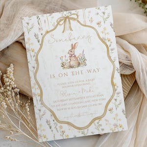 May include: Baby shower invitation with a floral design and a watercolor illustration of a bunny. The invitation features the text "Somebunny is on the way" and event details. The invitation is decorated with a gold ribbon and floral accents.
