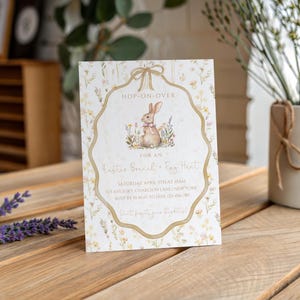 May include: An Easter-themed invitation card with a bunny illustration. The card features floral accents and the text "Hop-On-Over" and "Easter Bread & Egg Hunt". The invitation is on a wooden surface.