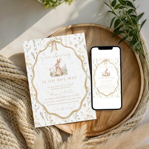 May include: A baby shower invitation with the text "Somebunny is on the way" and a watercolor illustration of a bunny. The invitation is on a wooden tray with a smartphone displaying the same design. The design includes floral elements and a bow.