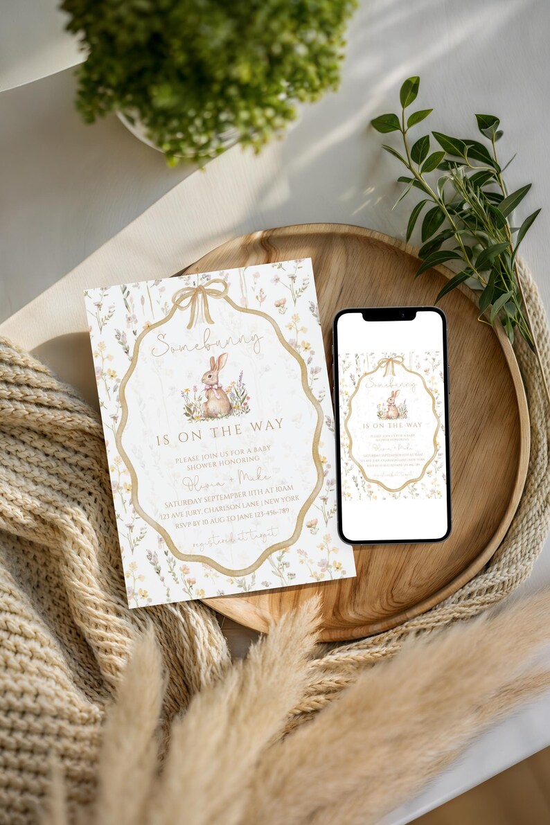 May include: A baby shower invitation with the text "Somebunny is on the way" and a watercolor illustration of a bunny. The invitation is on a wooden tray with a smartphone displaying the same design. The design includes floral elements and a bow.
