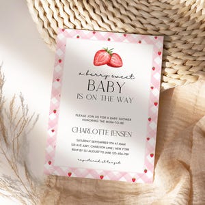 May include: A baby shower invitation with a pink gingham border and strawberry accents. The text reads "a berry sweet BABY IS ON THE WAY" with details for Charlotte Jensen's baby shower.