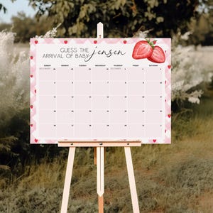 May include: A pink and white baby shower calendar on a wooden easel. The calendar has a strawberry theme with the text "Guess the Arrival of Baby Jensen". The calendar is decorated with strawberry illustrations.